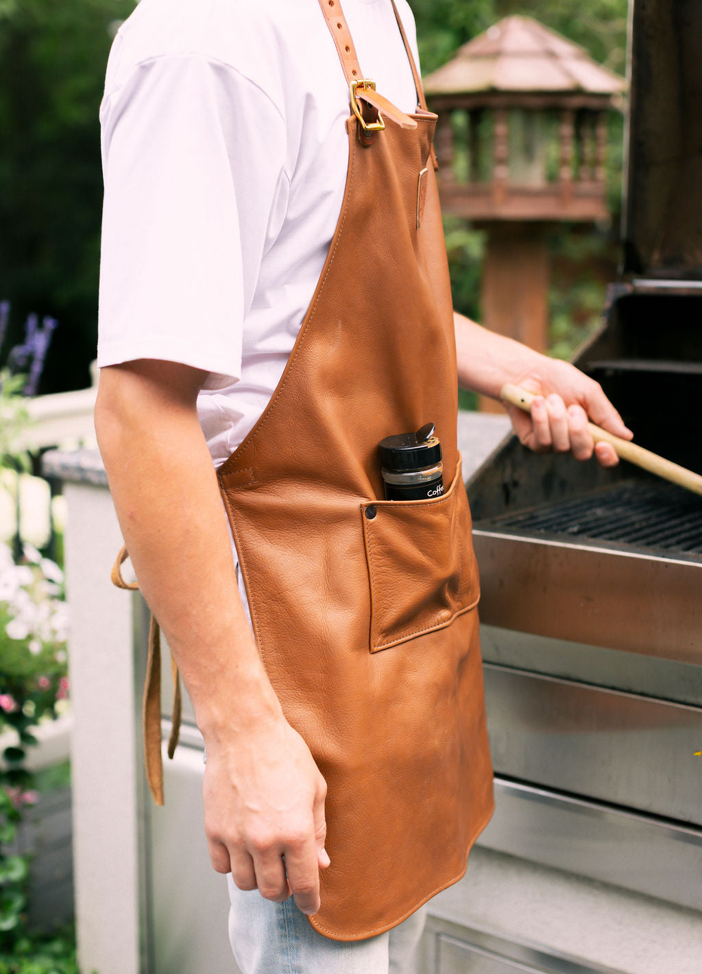 man wearing a brown leather apron outdoors near a grill, a seasoning bottle in the front pocket