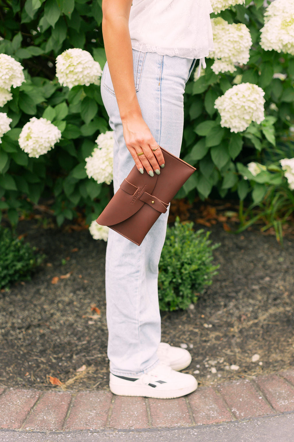 Woman holds a dark brown leather clutch standing in front of white flowers