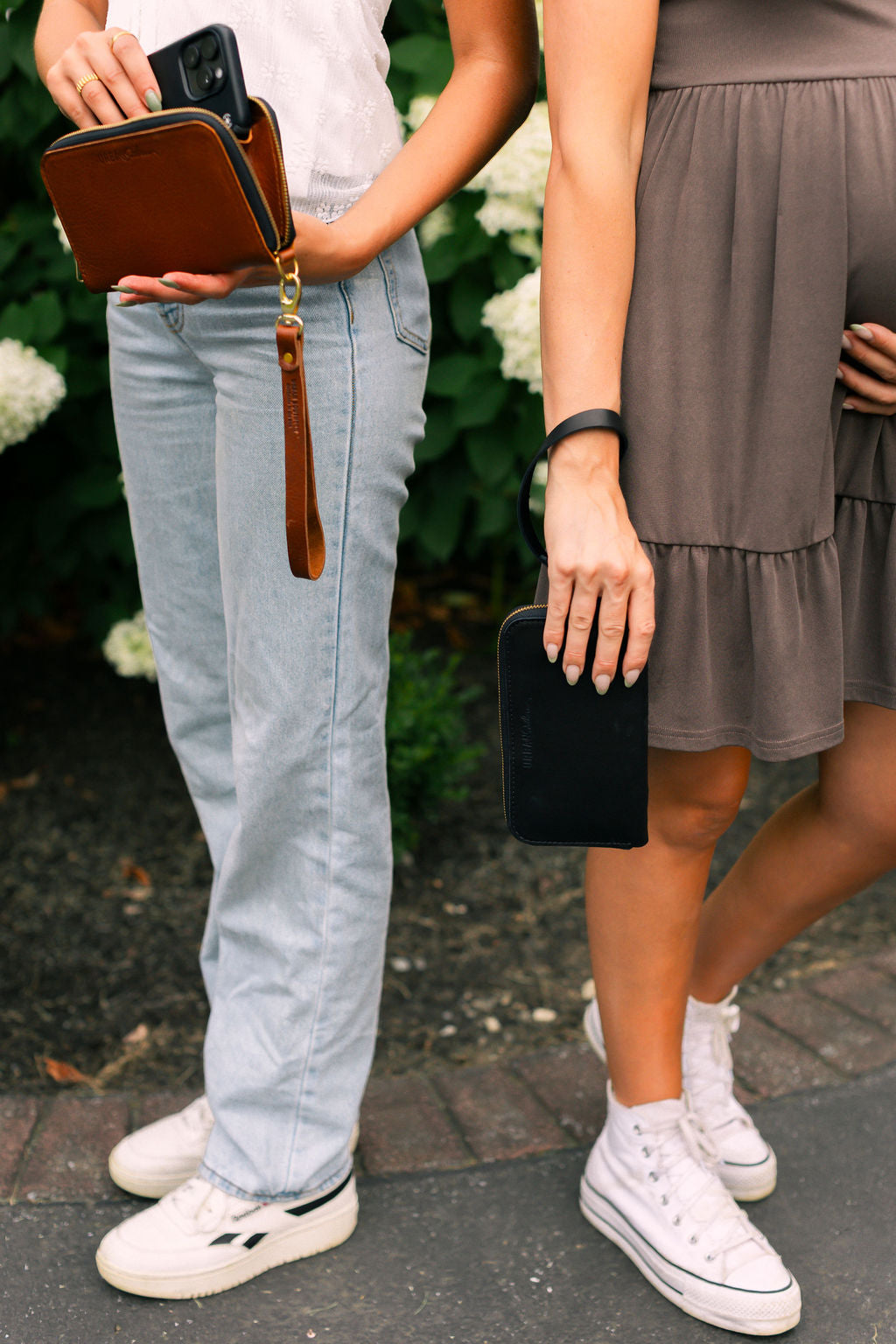 Two women stand in front of white flowers, one holds a brown leather wristlet wallet, one holds a black leather wristlet wallet. 