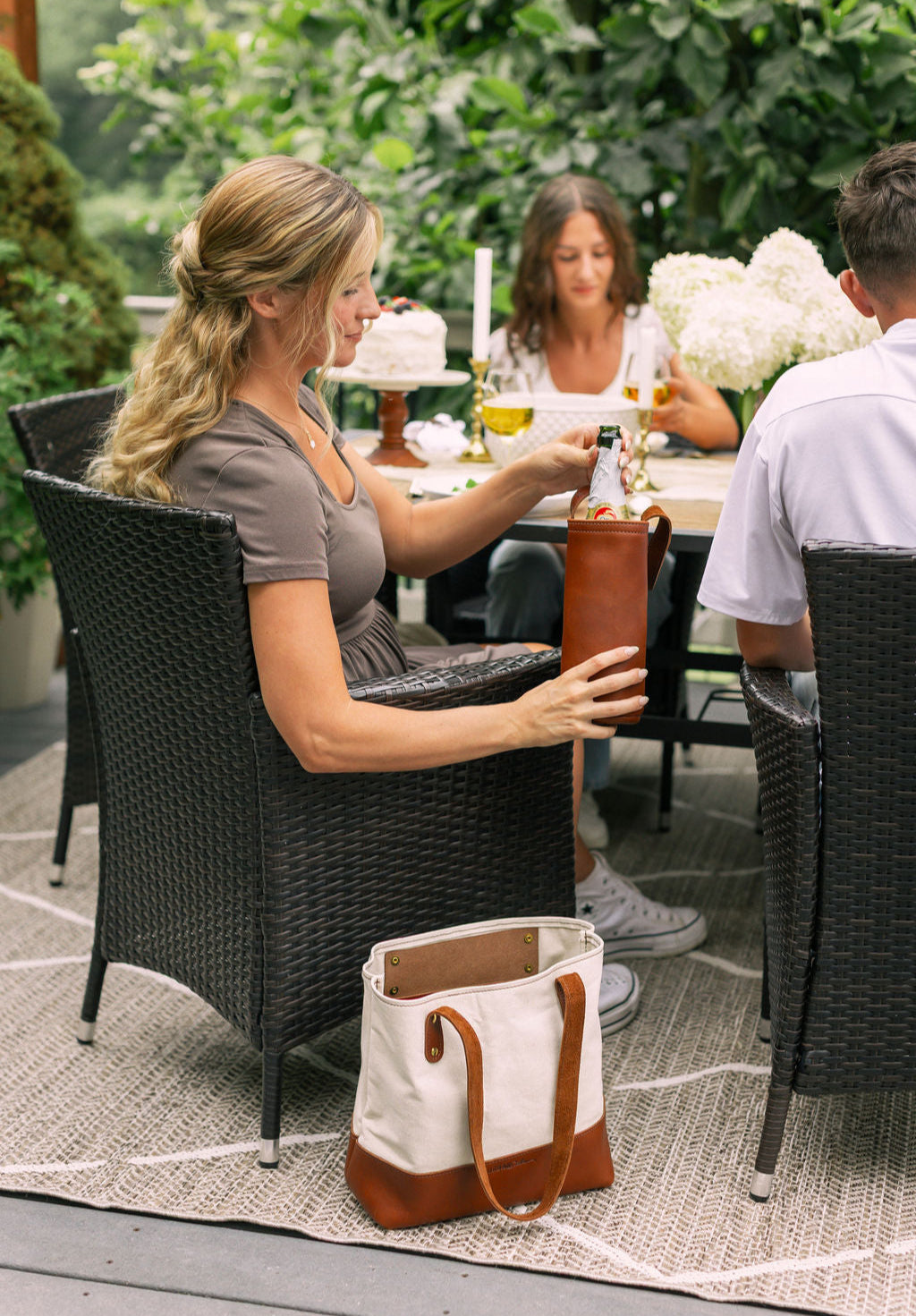 A woman sitting at an outdoor dining table, removes a bottle of sparkling cider from a leather wine tote holder, a handbag sits on the floor next to her.
