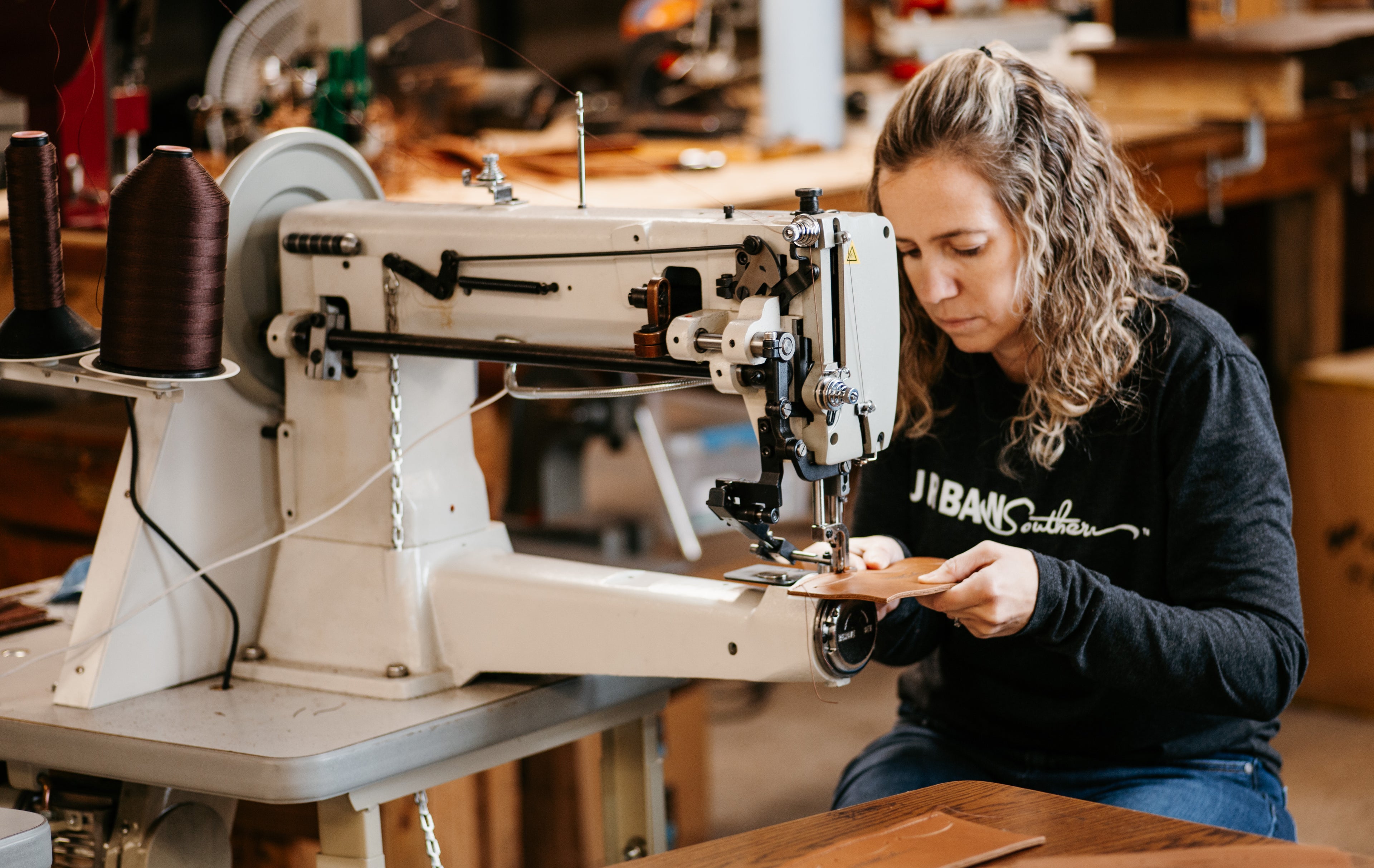 A woman sits at a sewing machine, sewing a leather handbag. 