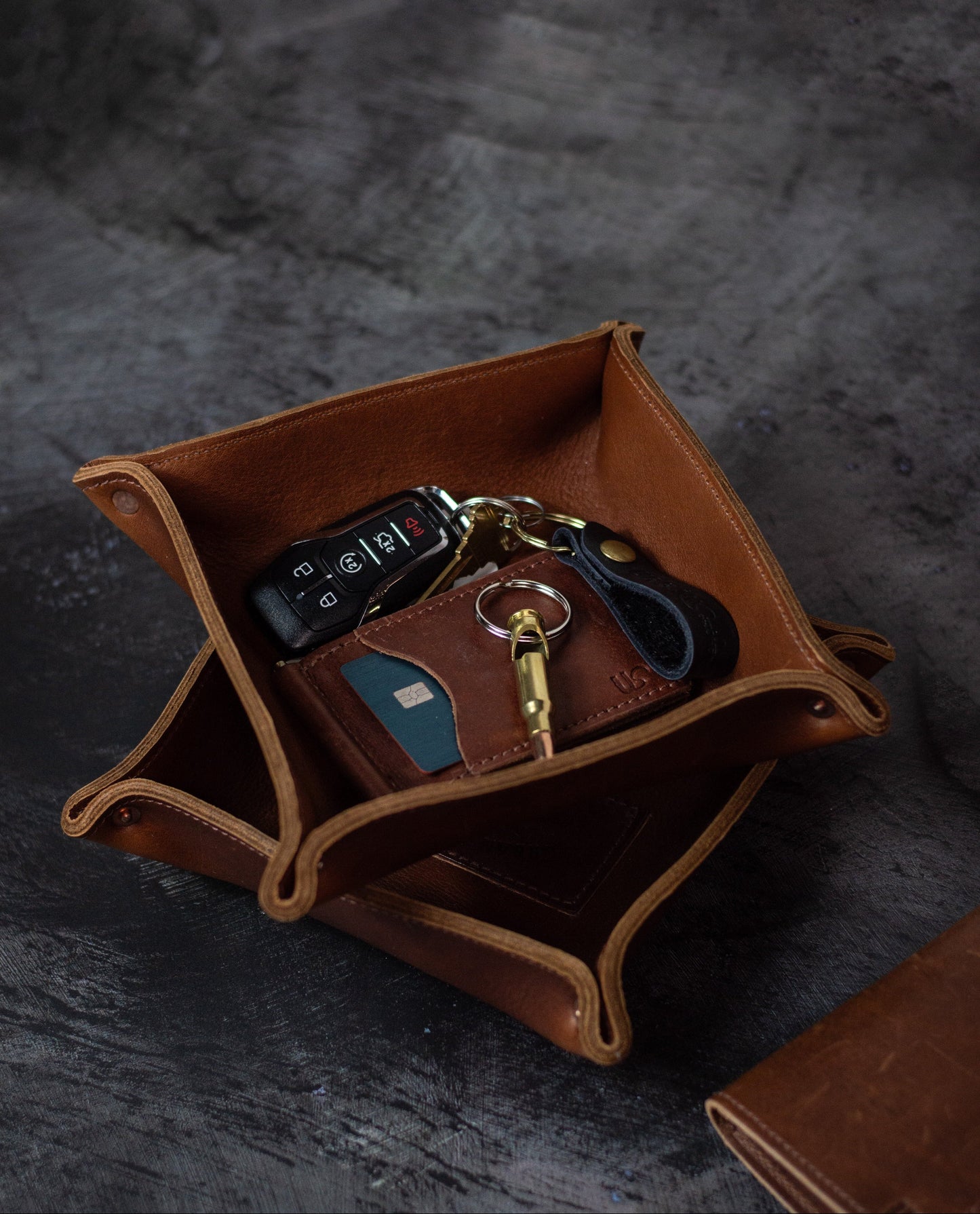Two leather valet trays sit on top of each other, one filled with a wallet & keys. On a black distressed background. 