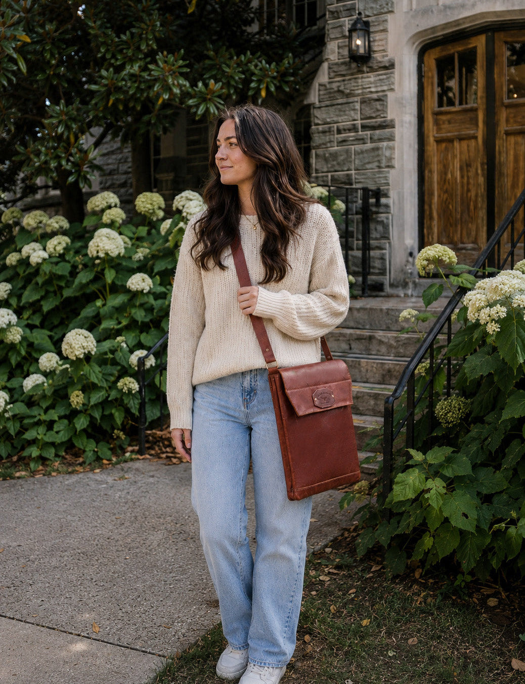 A woman stands on a stone porch walkway wearing a cream cable-knit sweater, light-wash blue jeans, white sneakers, and carrying a brown leather crossbody bag.