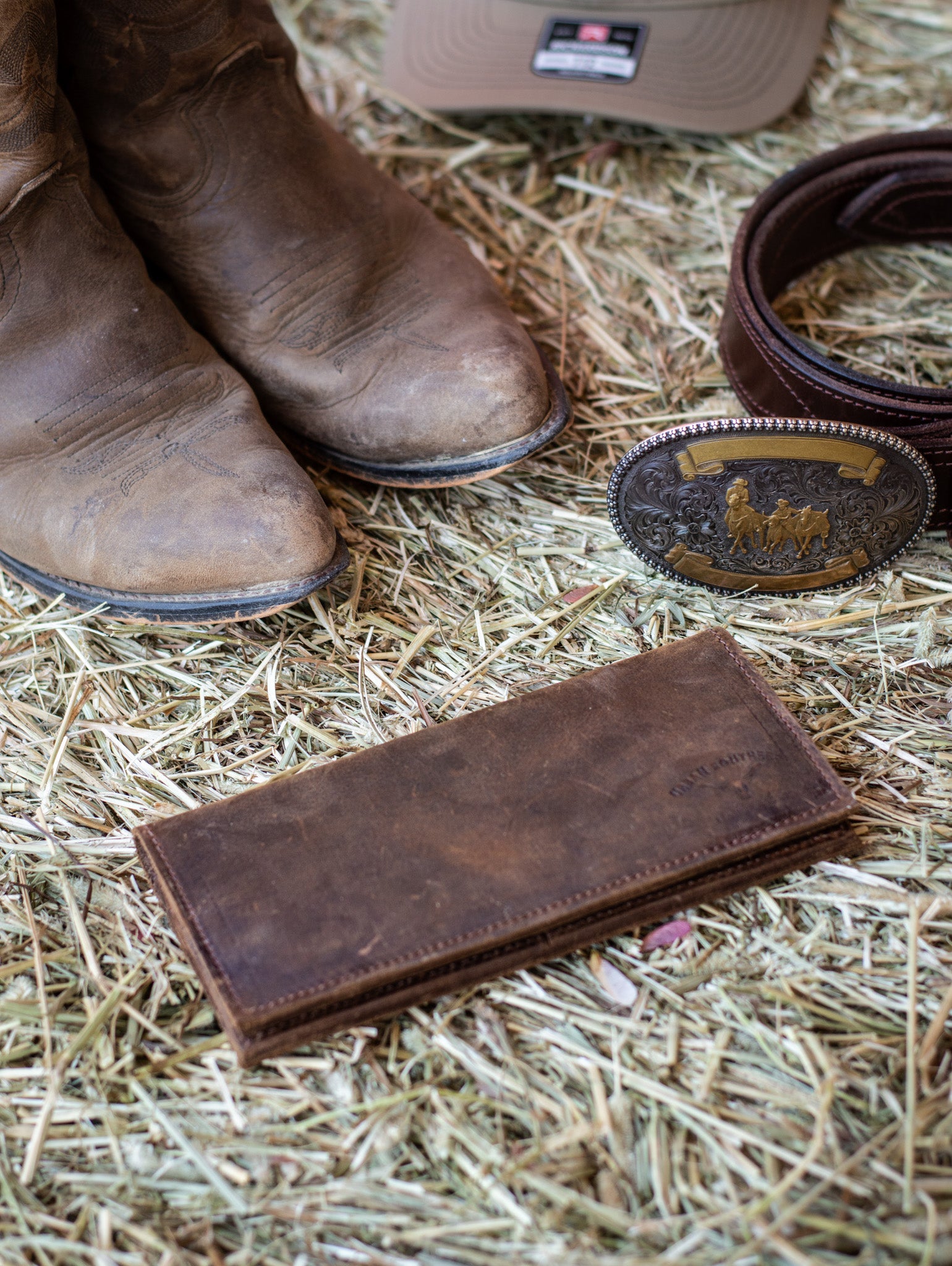 A rustic brown leather wallet, lies closed on a bale of hay next to a pair of cowboy boots and western belt buckle. 