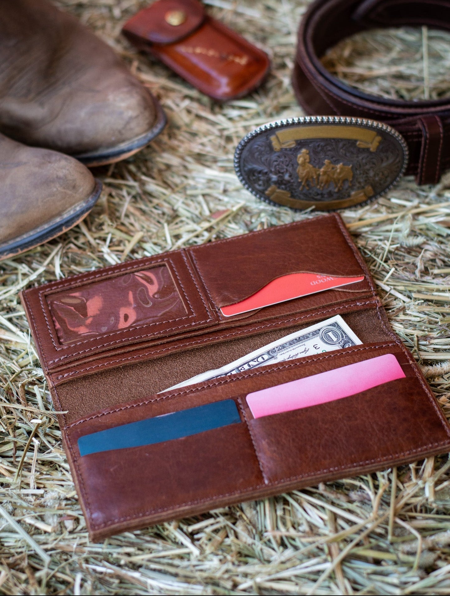A brown leather western wallet lies open on a hay bale, revealing credit cards within its card pockets & cash in its large slot. 