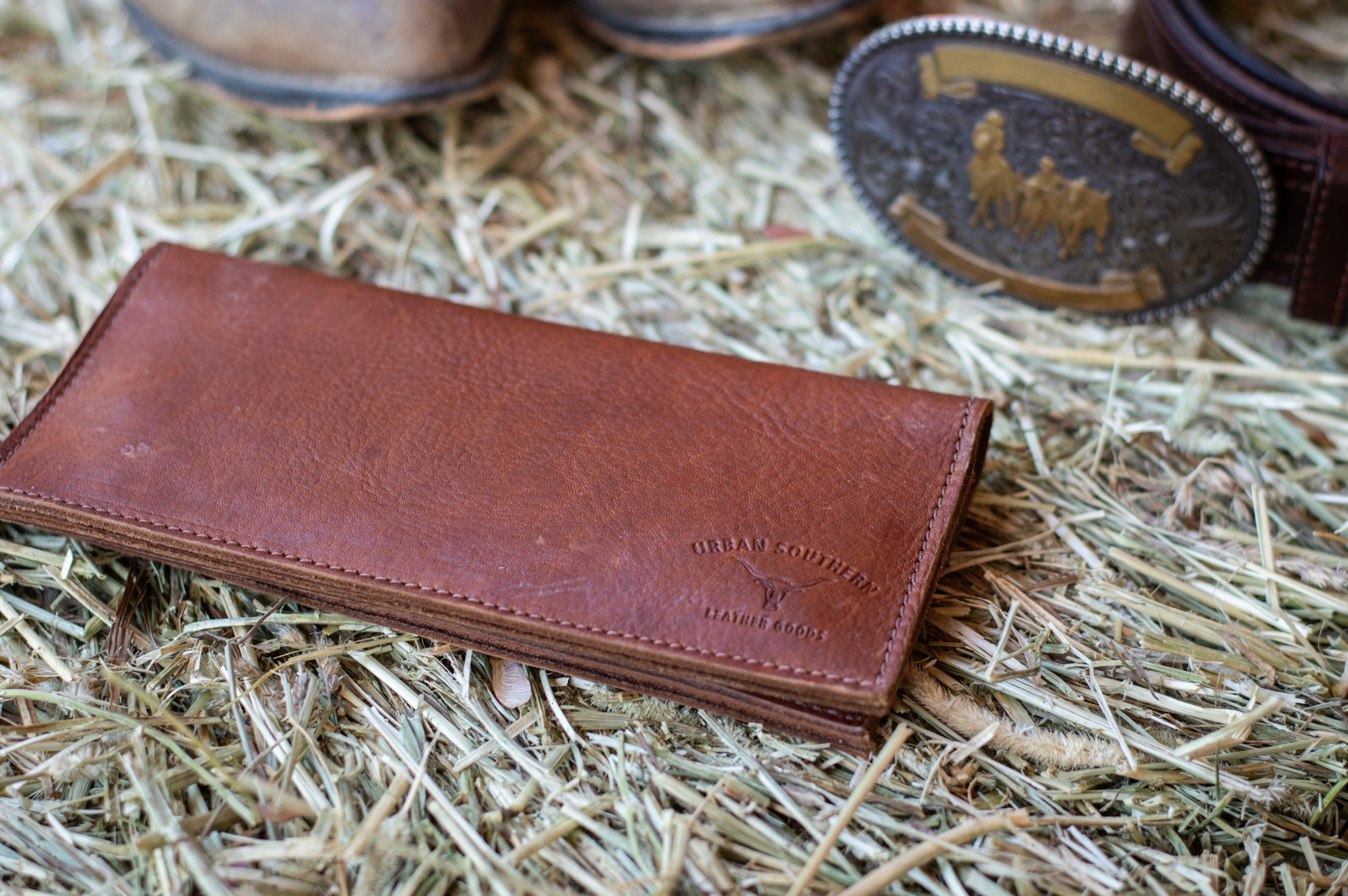 Cognac brown leather western wallet lies on a hay bale next to a western belt buckle. A brand on the corner of the wallet with the name 'urban southern' surrounds a longhorn steer design. 