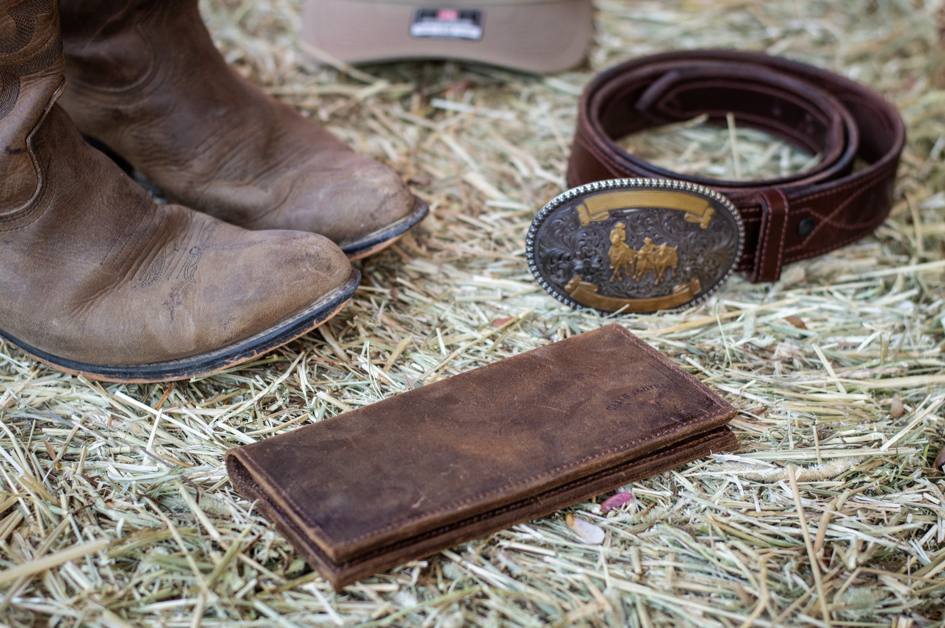 A rustic brown leather western wallet lies closed next to a pair of cowboy boots & leather belt.