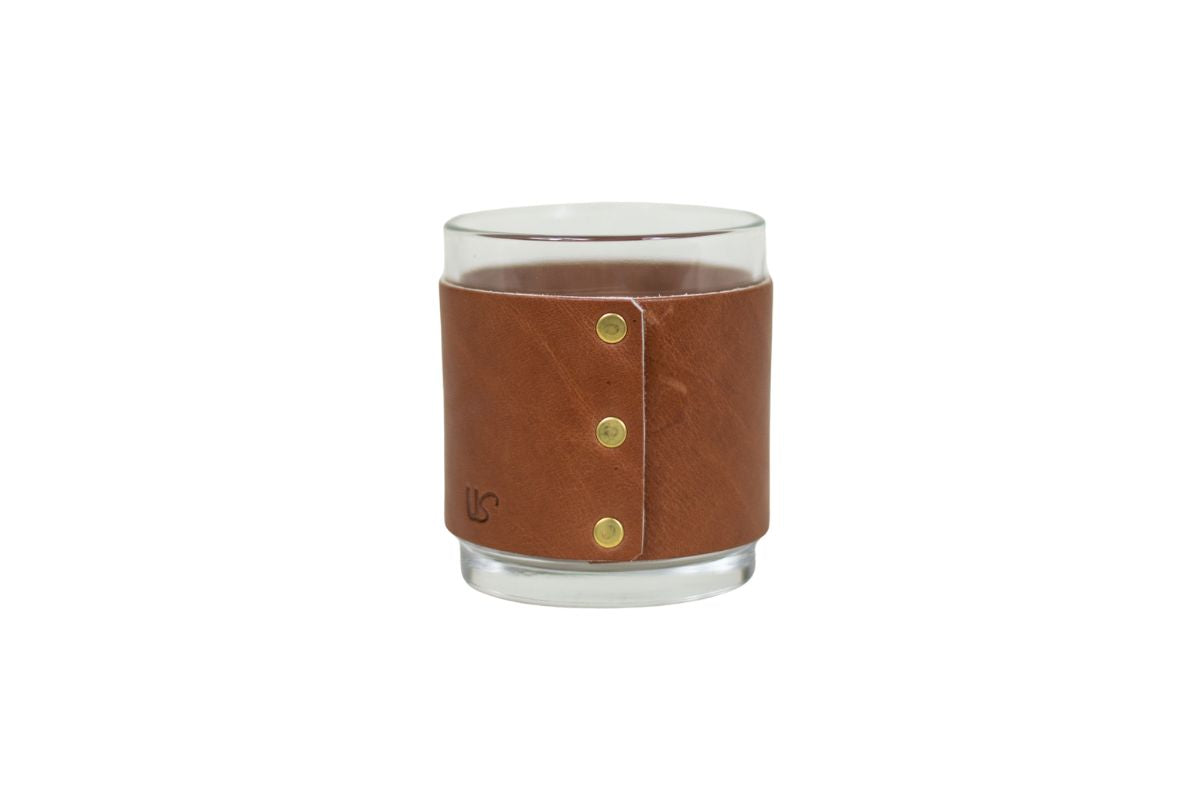 Clear whiskey glass with a brown leather sleeve featuring gold rivets on a white background