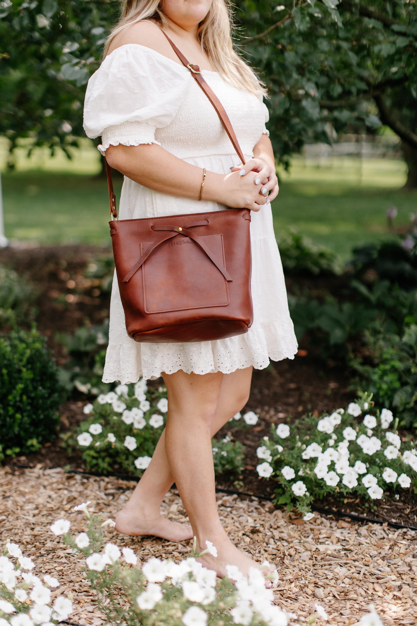 A woman walking down a garden path, holds a Cognac Lexington Bow handbag on her shoulder. 