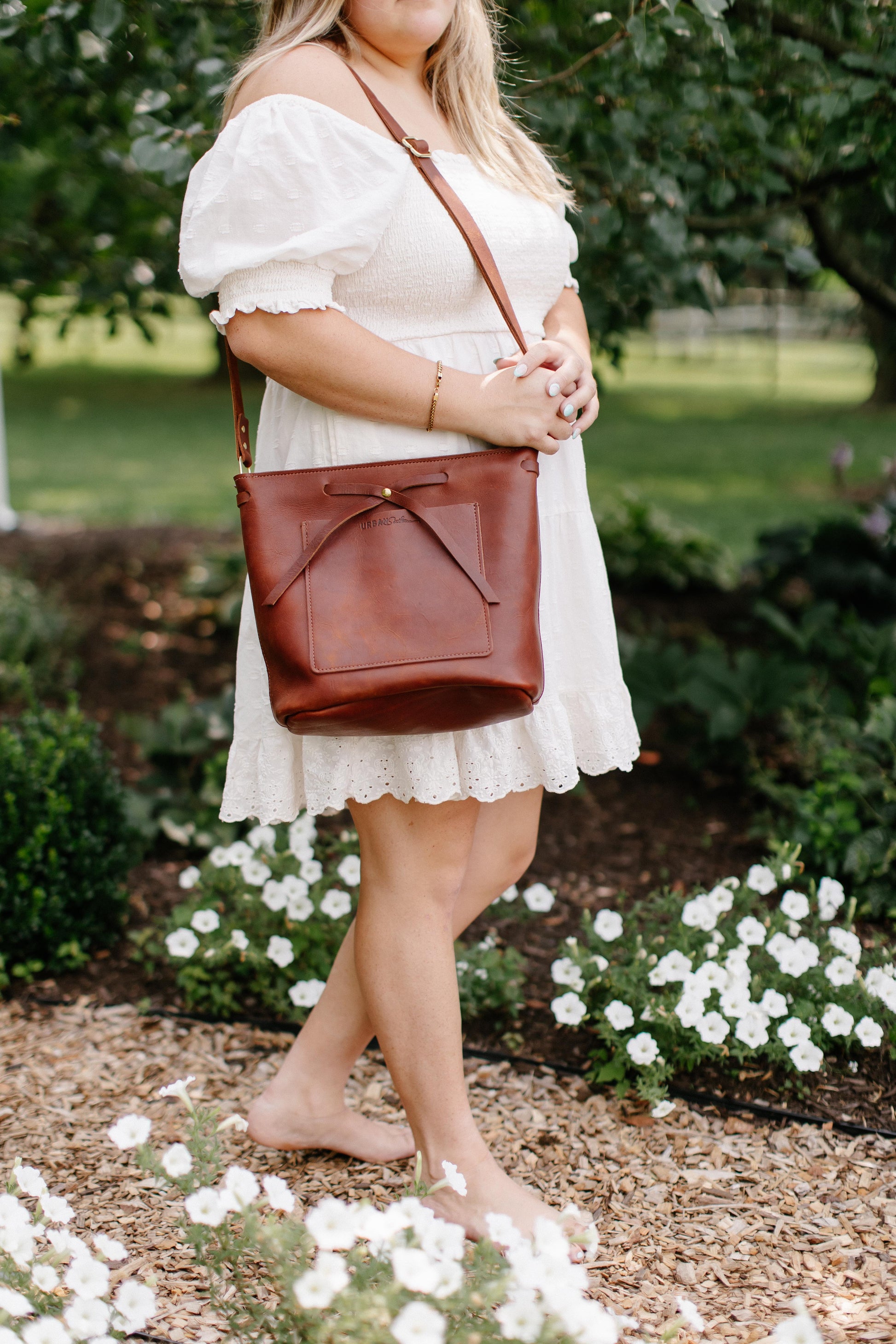 A woman walking down a garden path, holds a Cognac Lexington Bow handbag on her shoulder. 