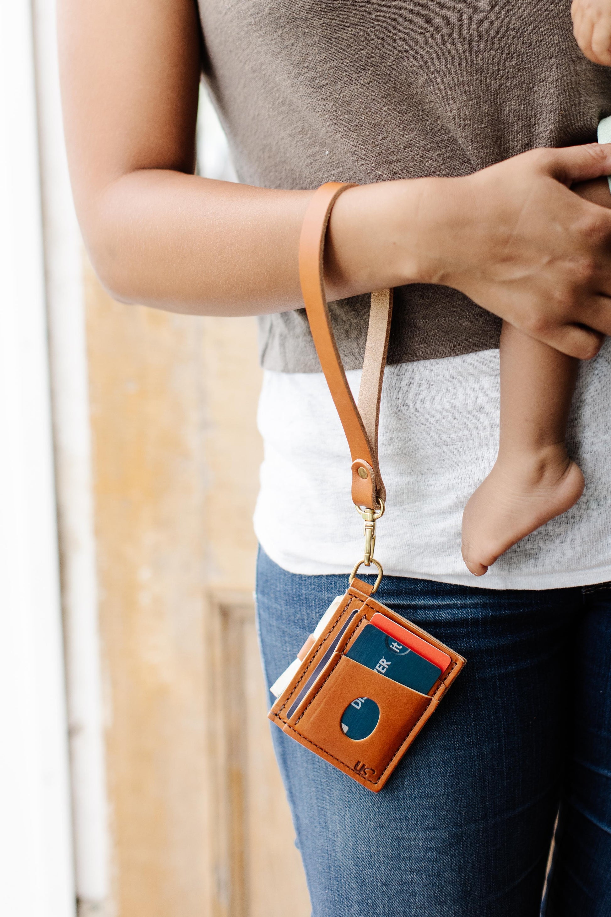 The Urban Southern handcrafted leather Everly Wristlet Wallet in Tan. 
A woman carries the Everly Wristlet Wallet in Tan on her wrist while carrying a child.