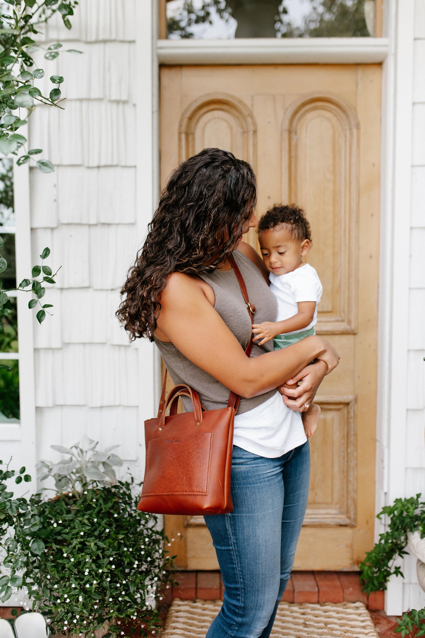A woman holds a small child in her arms while wearing a cognac Quinn Crossbody across her body. 