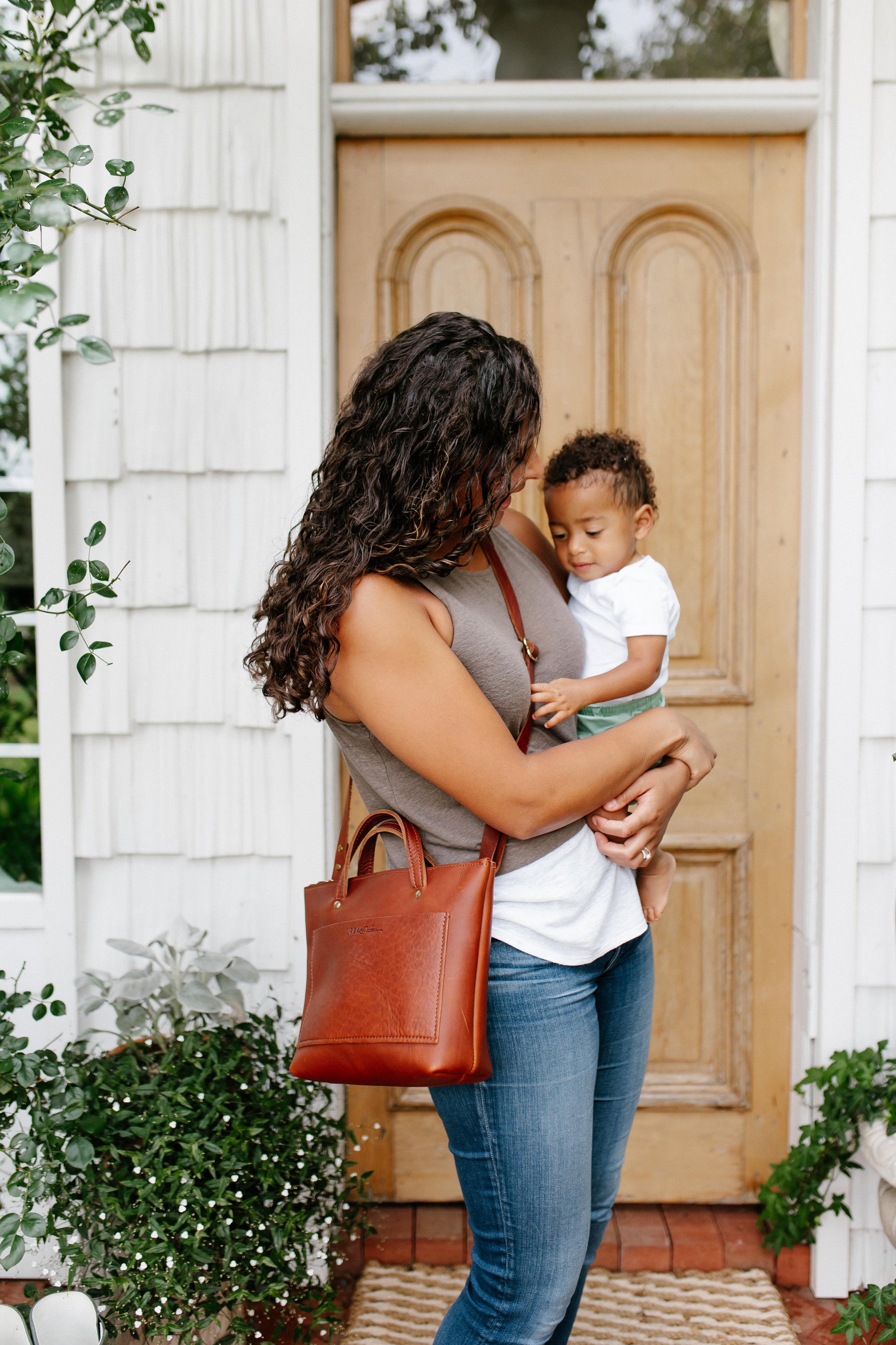 A woman holds a small child in her arms while wearing a cognac Quinn Crossbody across her body. 