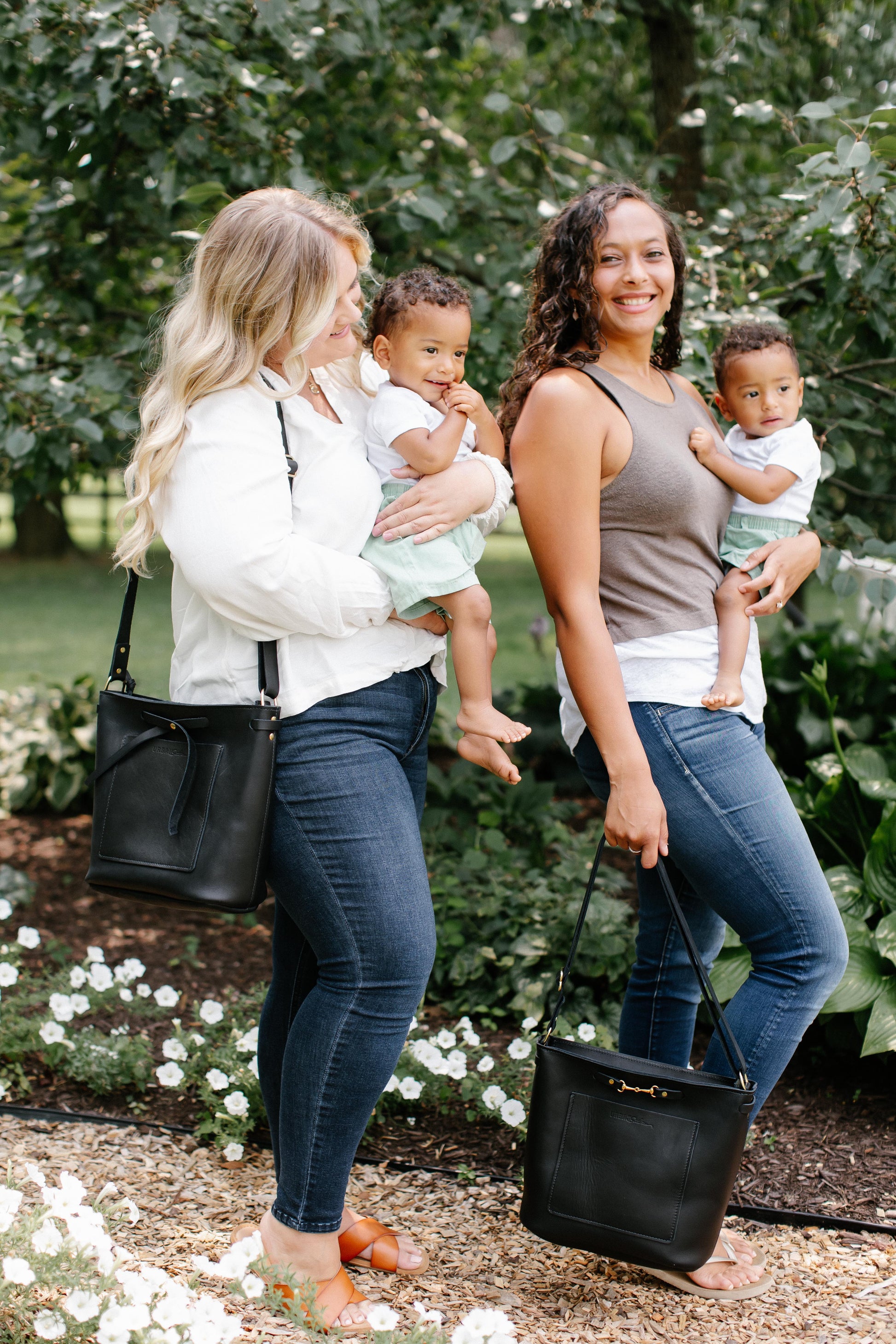 Two women walk through a garden path, each holding a small child. One holds a Black Lexington Julep handbag in her hand, while the other has a black Lexington Bow handbag on her shoulder. 