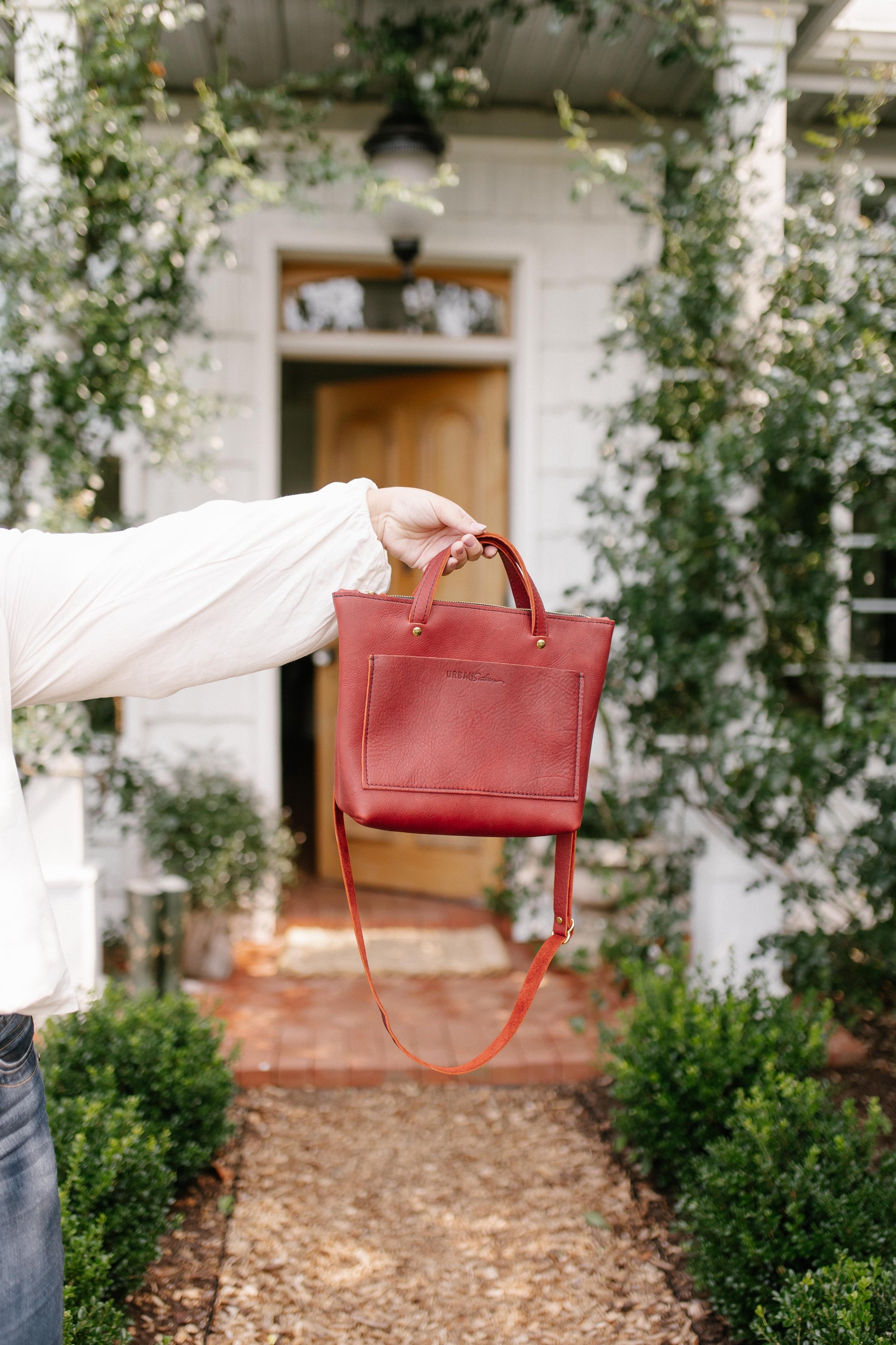 A person in a white long-sleeve top holds up a small red leather handbag with a matching shoulder strap, set against a blurred background of a white house and garden path.