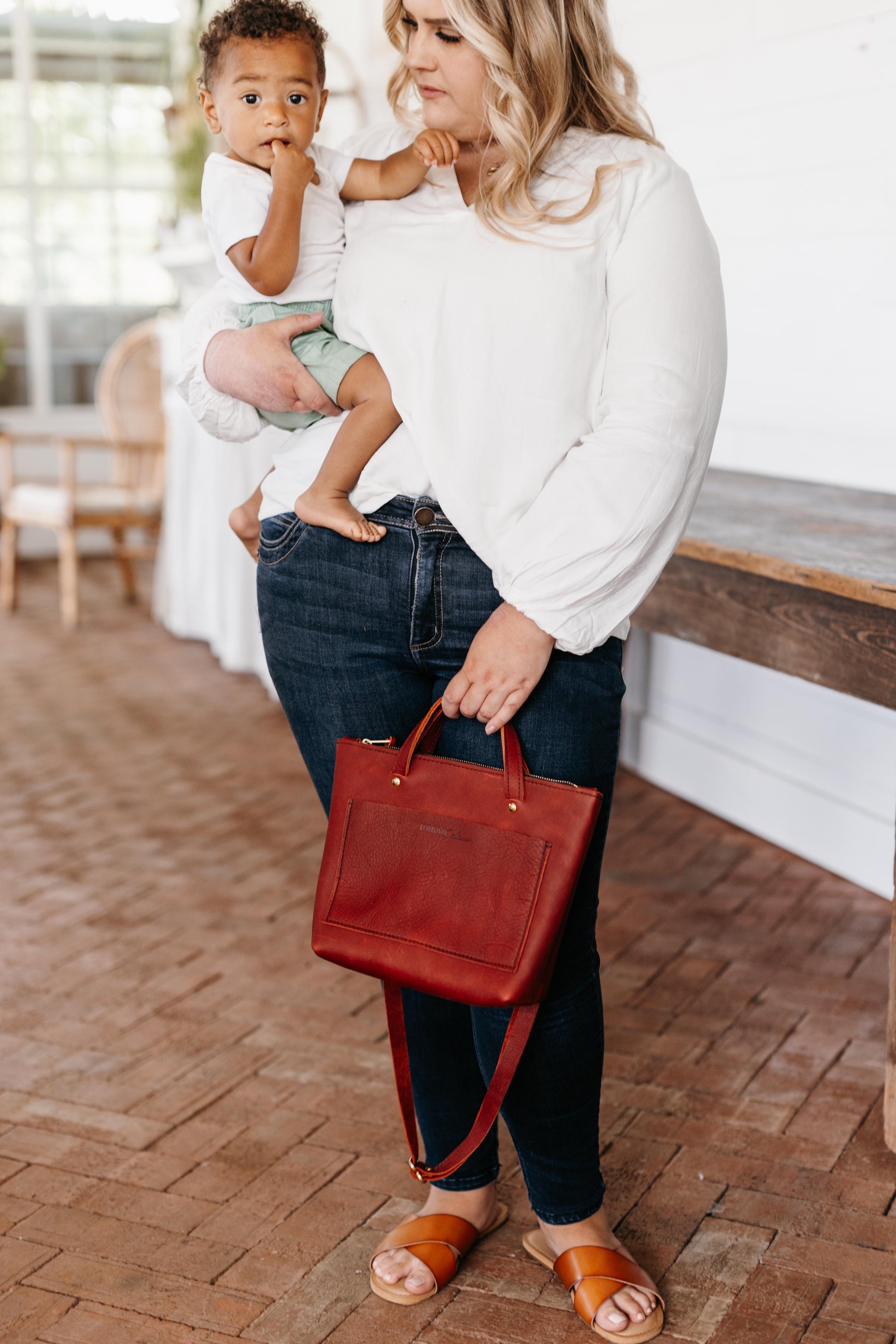 A woman holds a small child in her arm while holding a merlot Quinn Crossbody in her other hand by its convenient grab handles. 