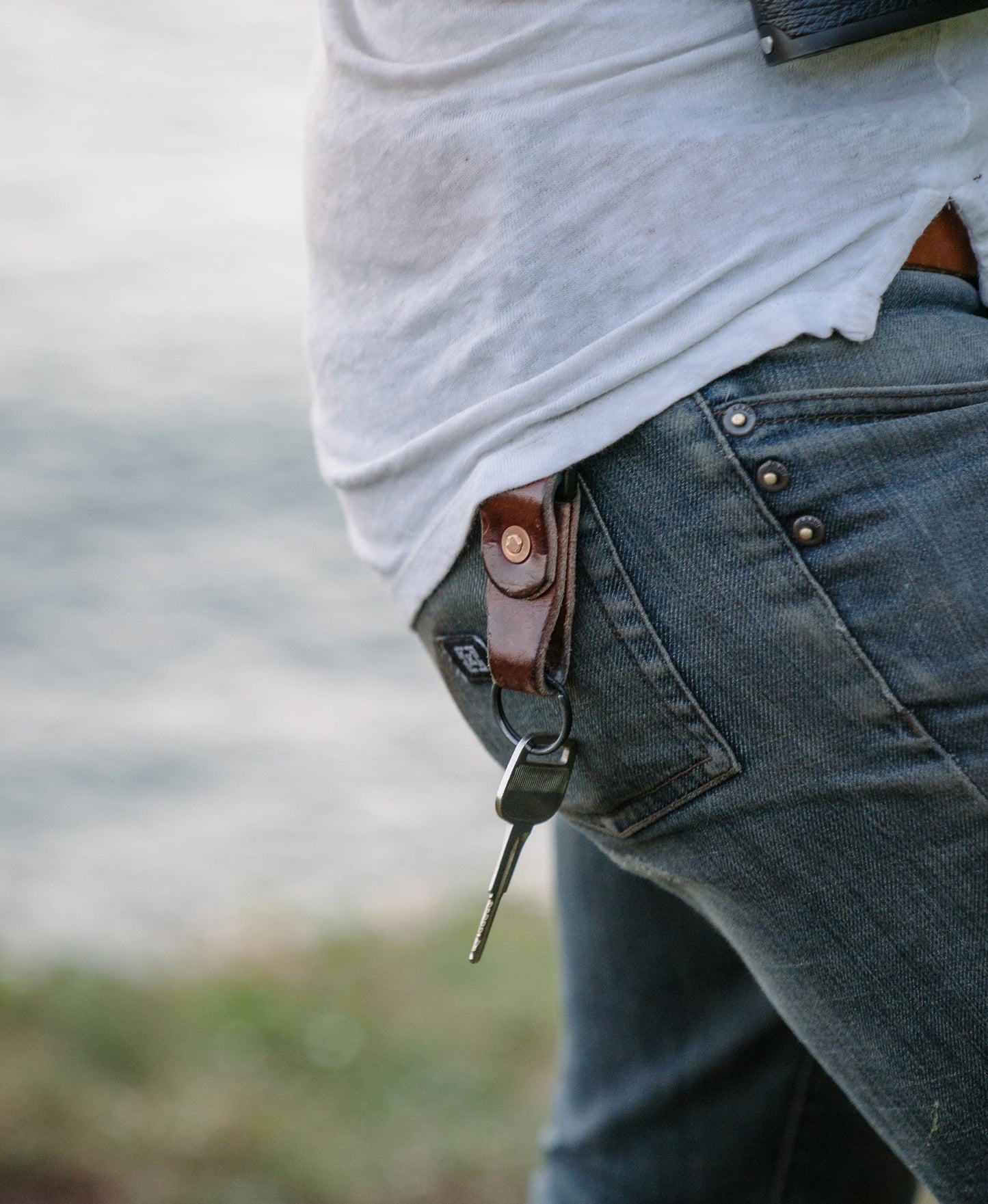 A man wearing a white shirt and jeans, has a leather keychain & set of keys clipped to his belt loop