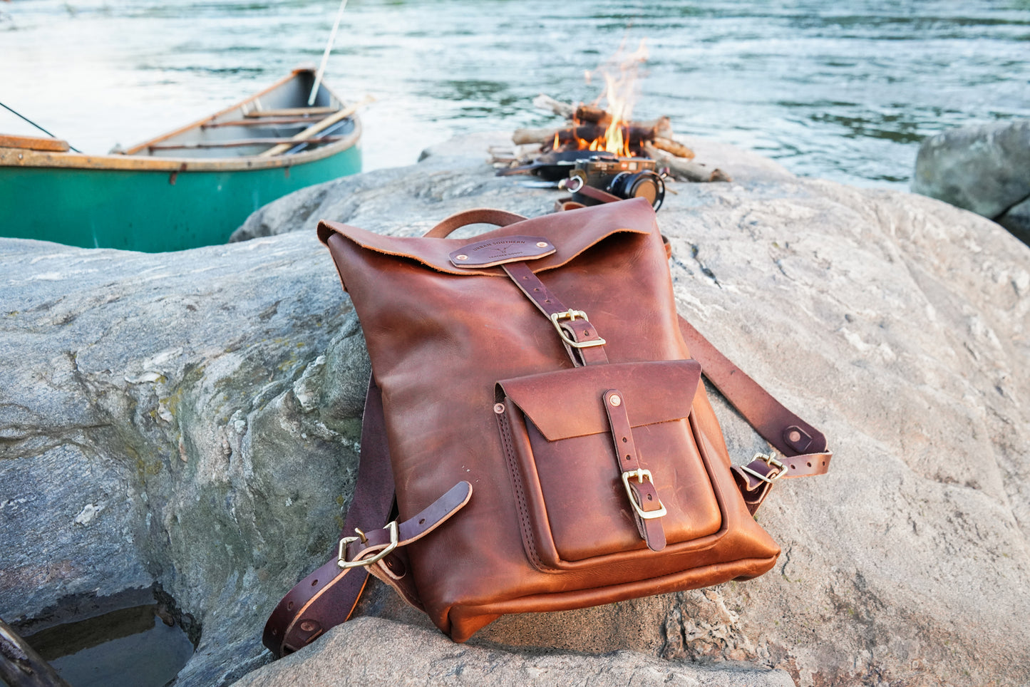 Brown leather backpack on a rock by a campfire and water