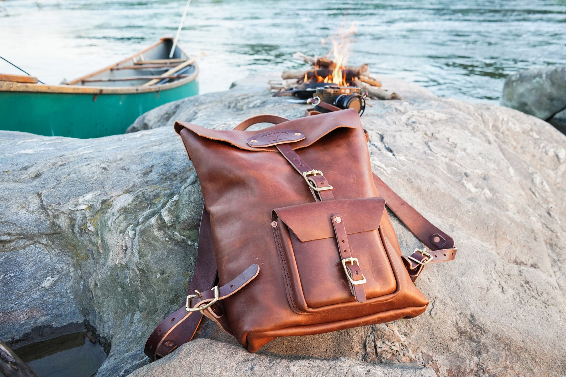 Brown leather backpack on a rock by a campfire and water