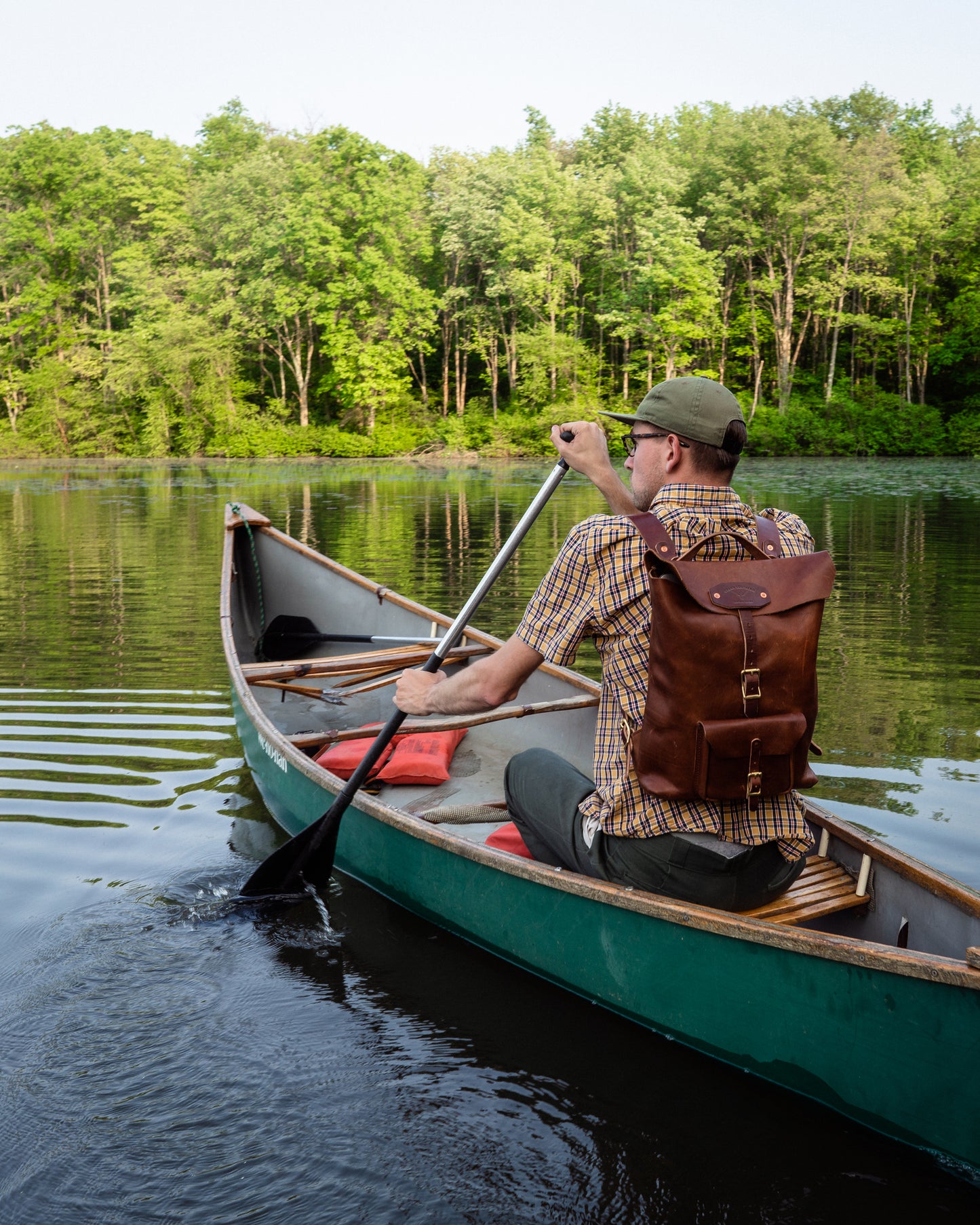 Man paddling a canoe on a calm lake with trees in the background, while wearing a brown leather rucksack on his back.
