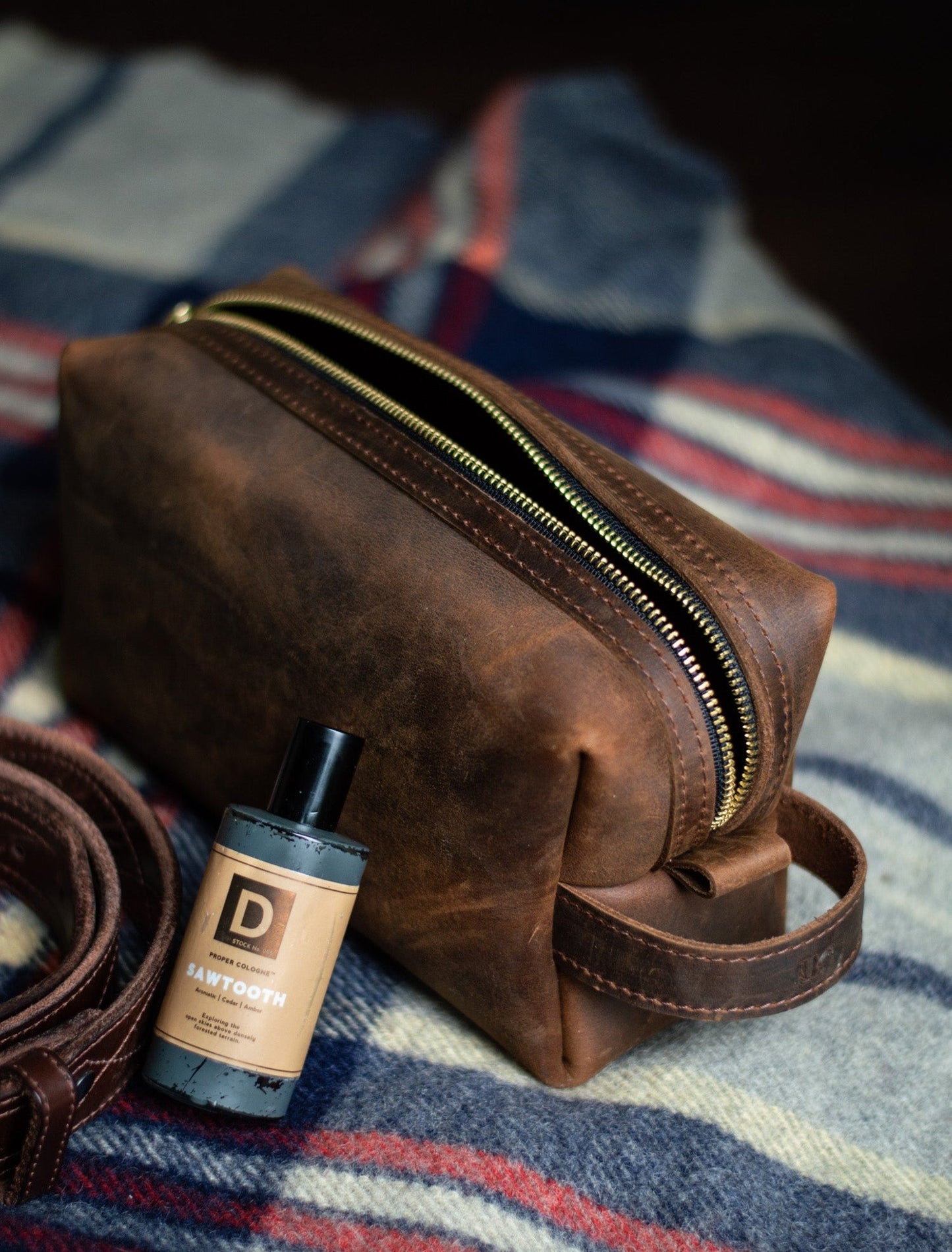 Rustic brown leather dopp kit with a bottle of aftershave on a plaid fabric background