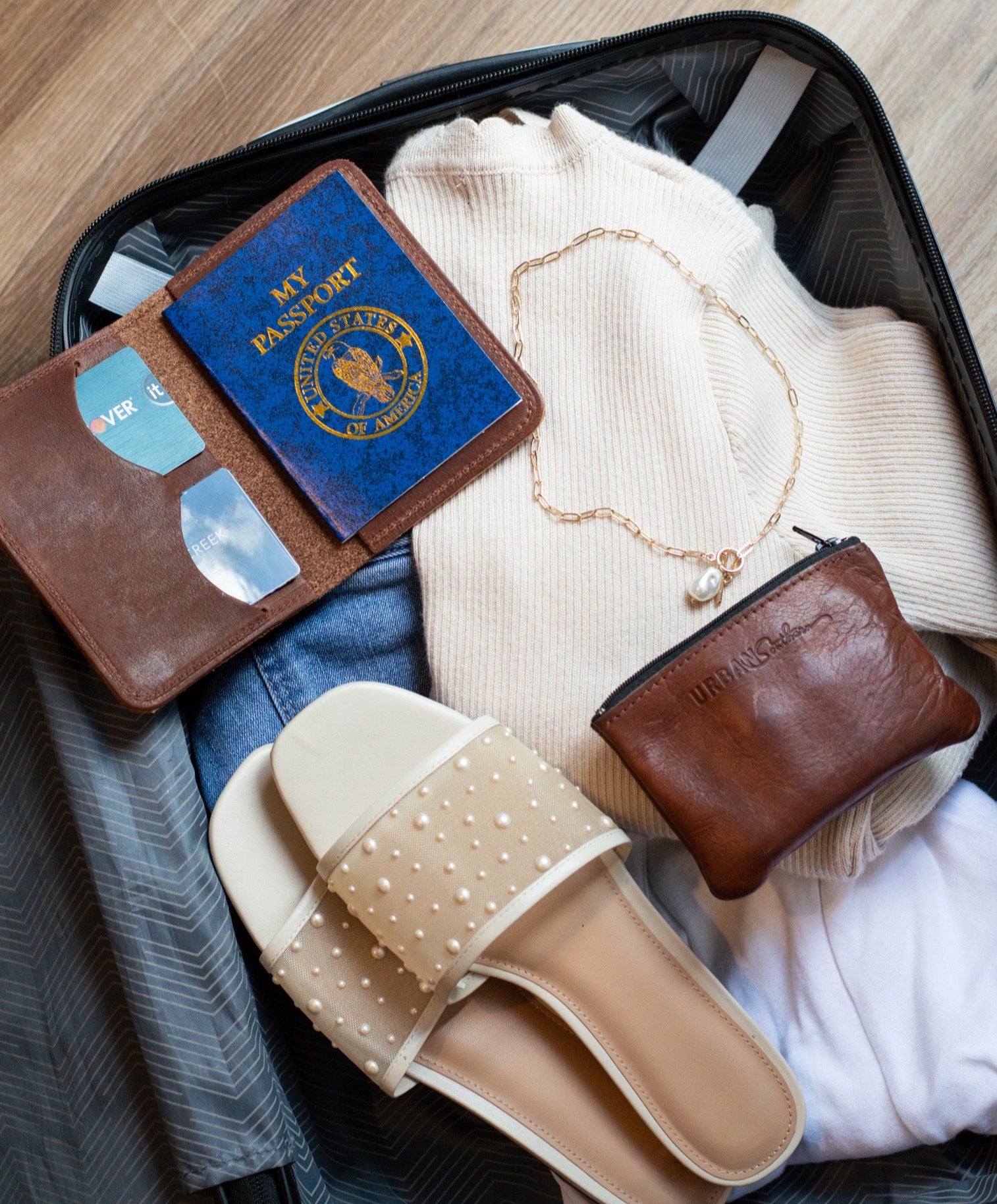 Open suitcase with travel items including a  leather passport wallet, jewelry, and clothing.