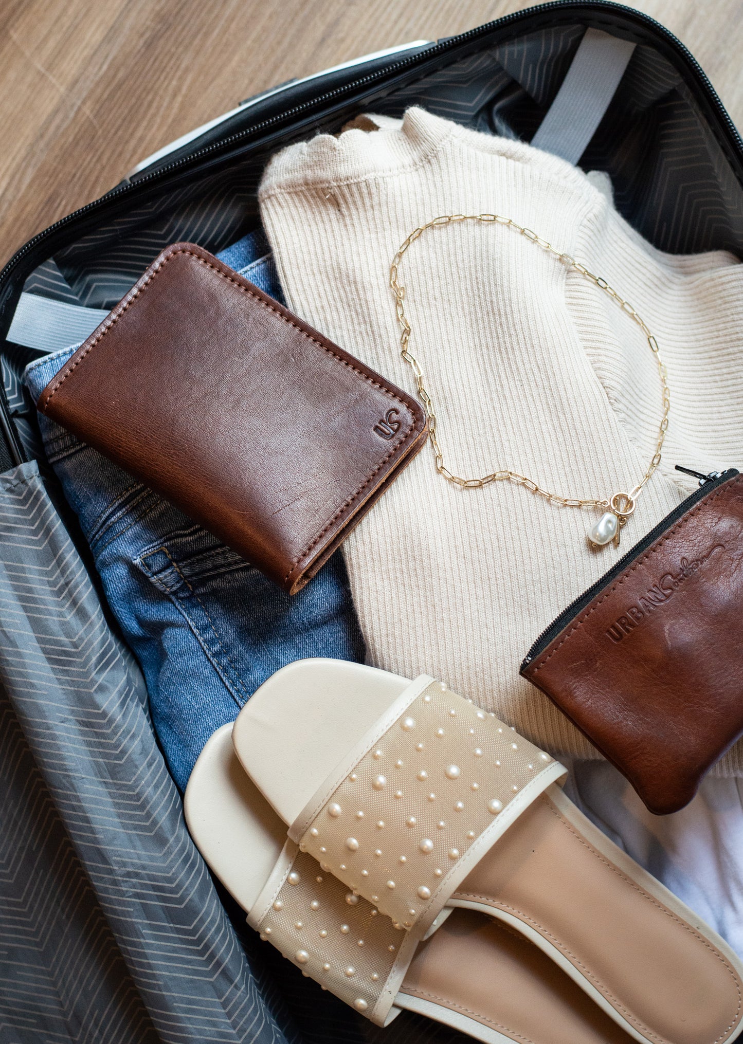 Brown passport wallet, necklace, beige sandals, and white sweater in an open suitcase on a wooden floor.