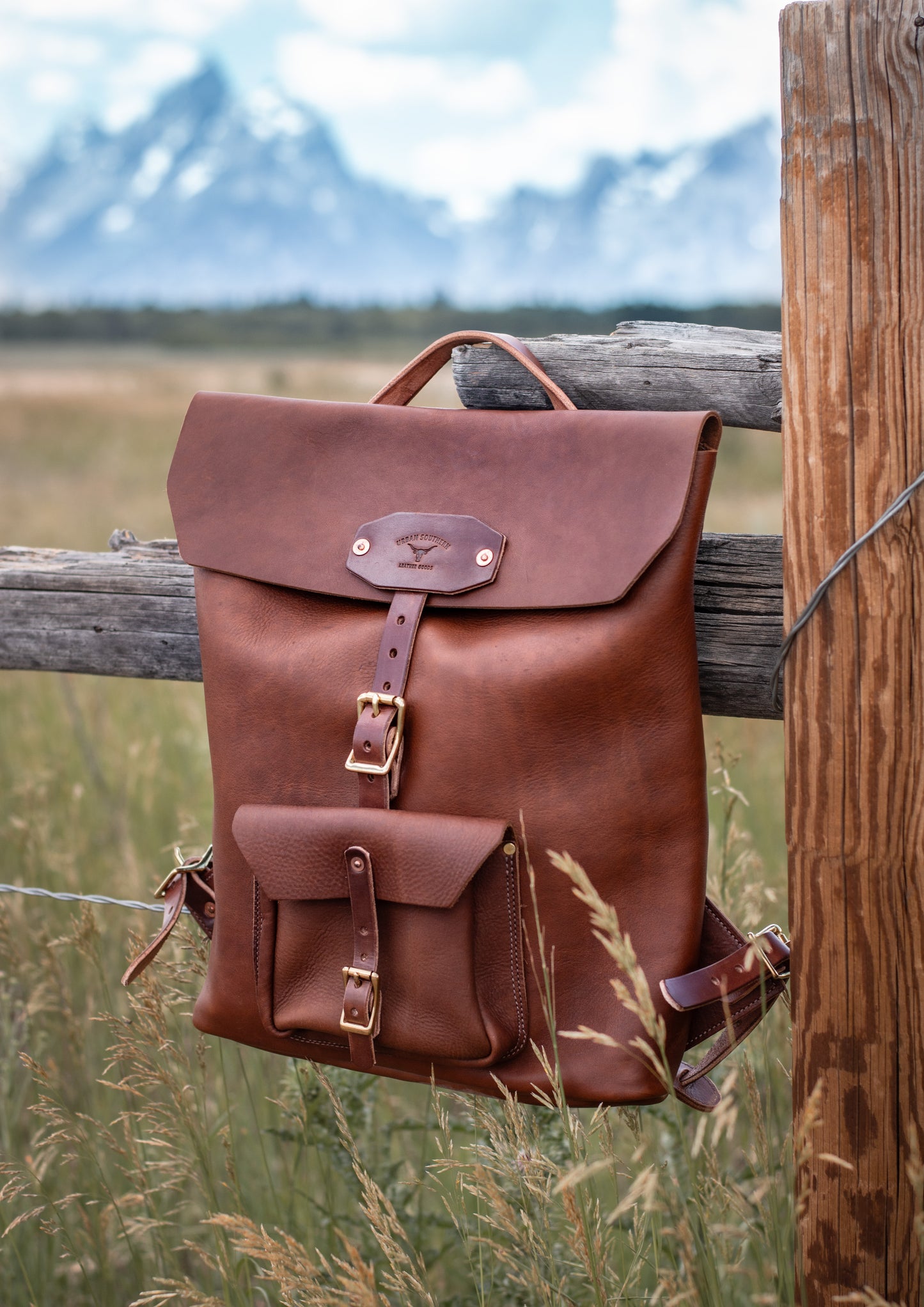 a brown leather backpack hangs on a wood fence with the Tetons mountain range on the background.