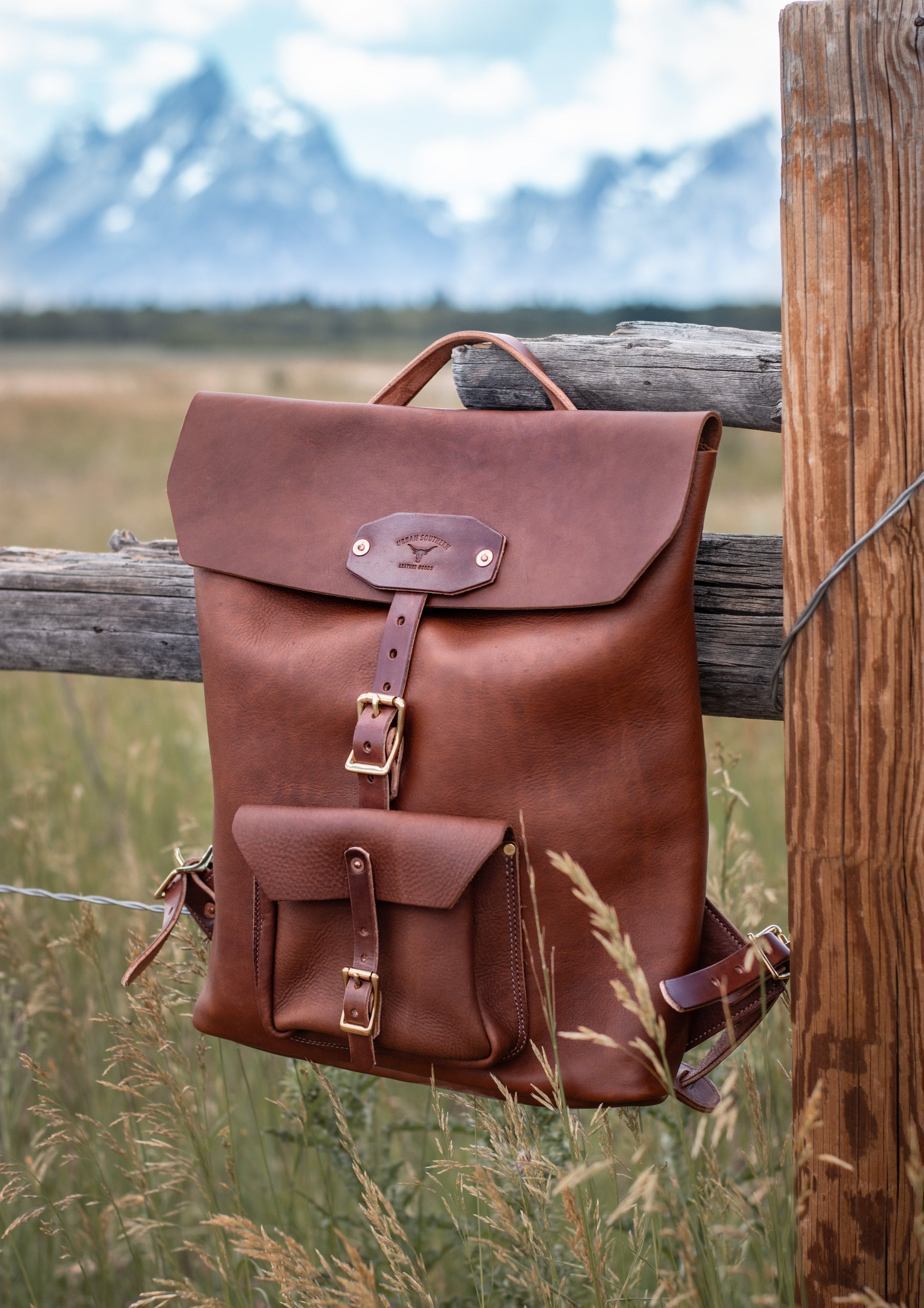 a brown leather backpack hangs on a wood fence with the Tetons mountain range on the background.