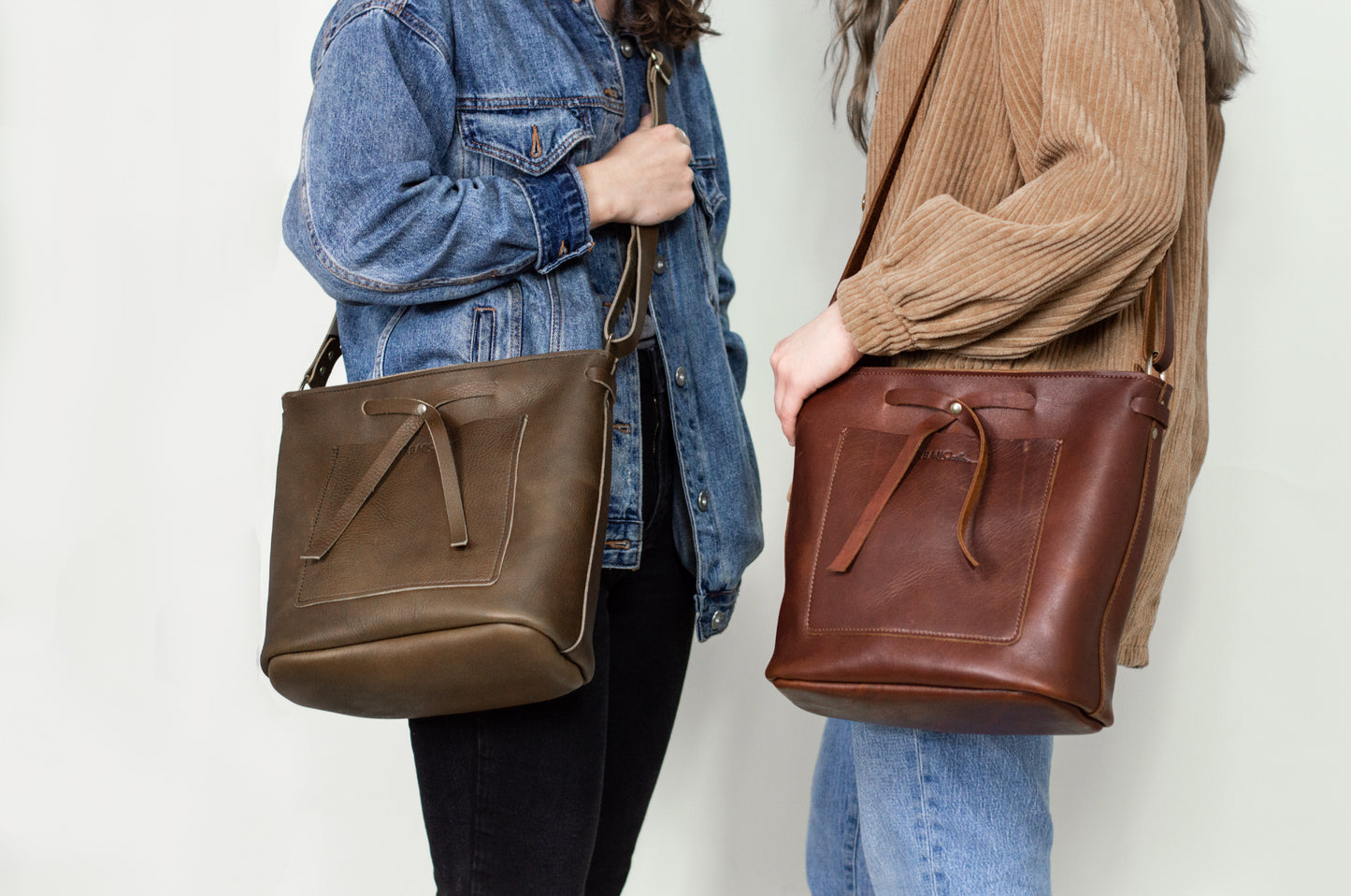 Two women stand near each other, one wearing a mocha Lexington bow handbag across her body, while the other wears a cognac Lexington Bow on her shoulder. 