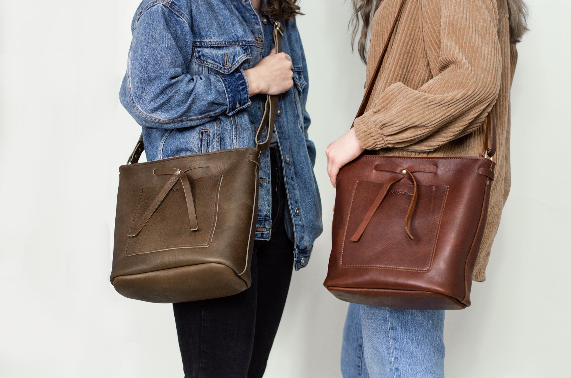 Two women stand near each other, one wearing a mocha Lexington bow handbag across her body, while the other wears a cognac Lexington Bow on her shoulder. 