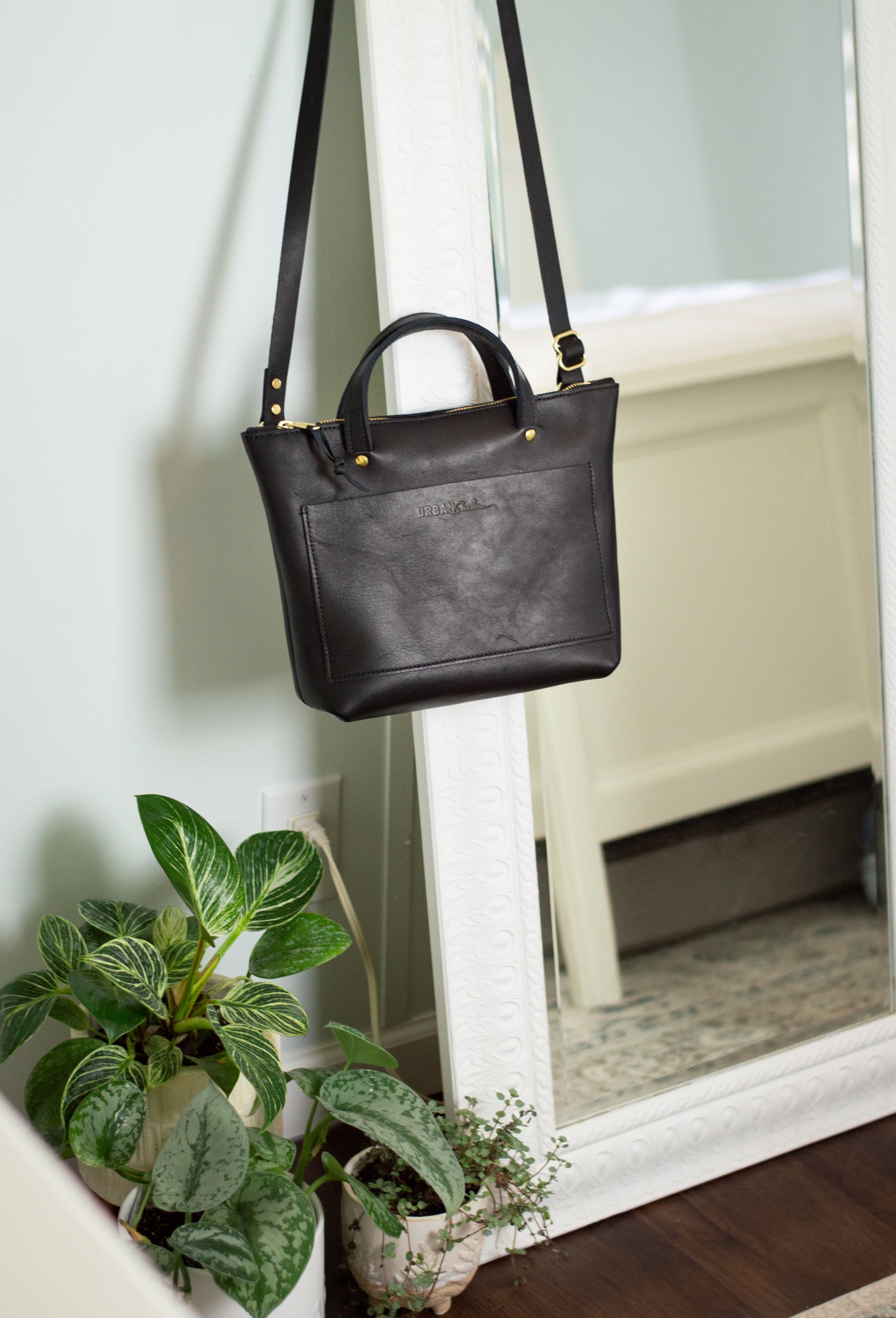 A black Quinn Crossbody hangs by its adjustable leather strap, on a mirror in a bedroom. 